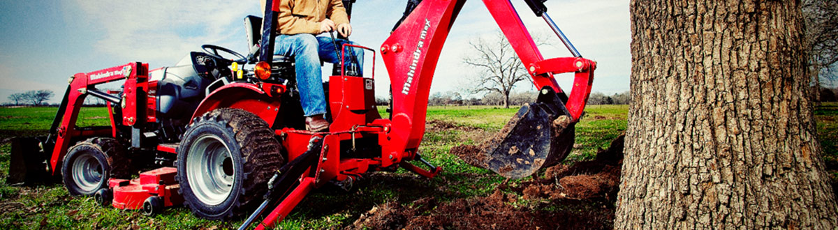 Person on a Mahindra® Backhoe Loader 1526B digging up the roots of a tree.